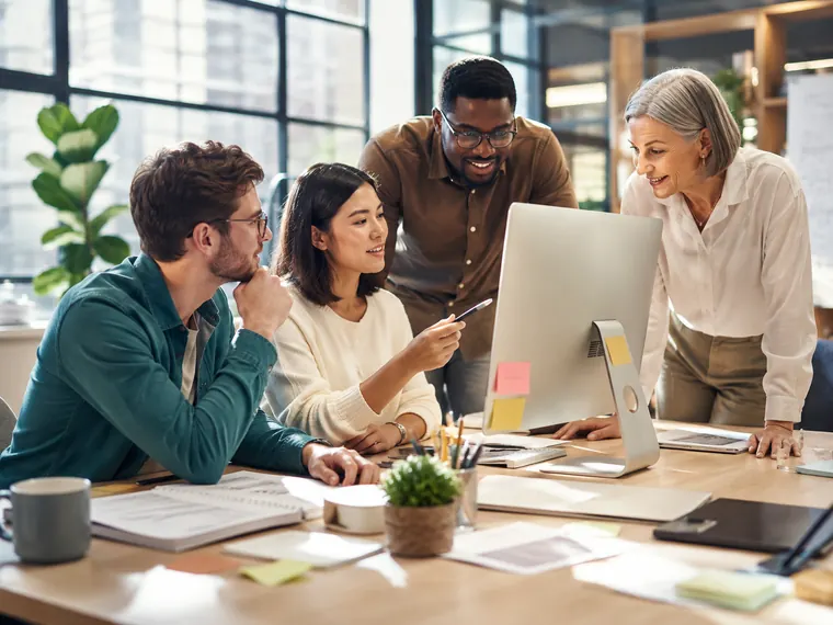 Four colleagues gathered around a desktop screen in a bright office, talking through their marketing together.